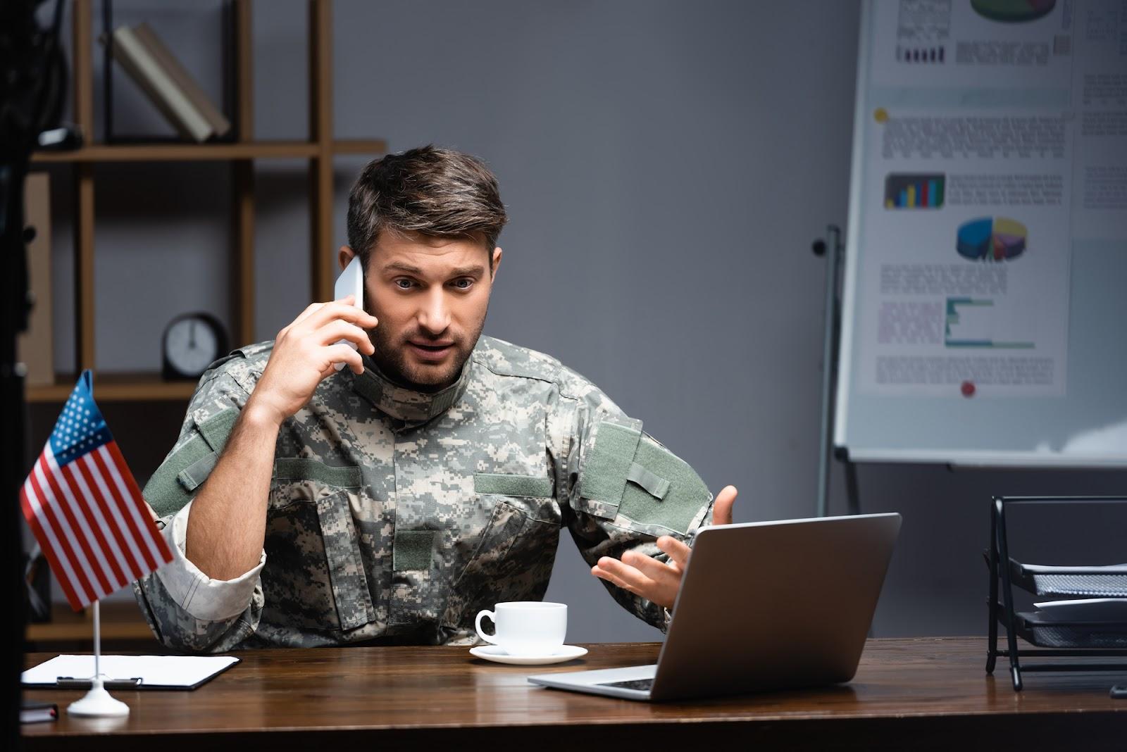 A man in military fatigues talking on a phone, sitting in front of an open laptop at a desk with a cup of coffee and an American flag, explaining why he cannot attend a court hearing; concept for requesting stay of proceedings in a military divorce.