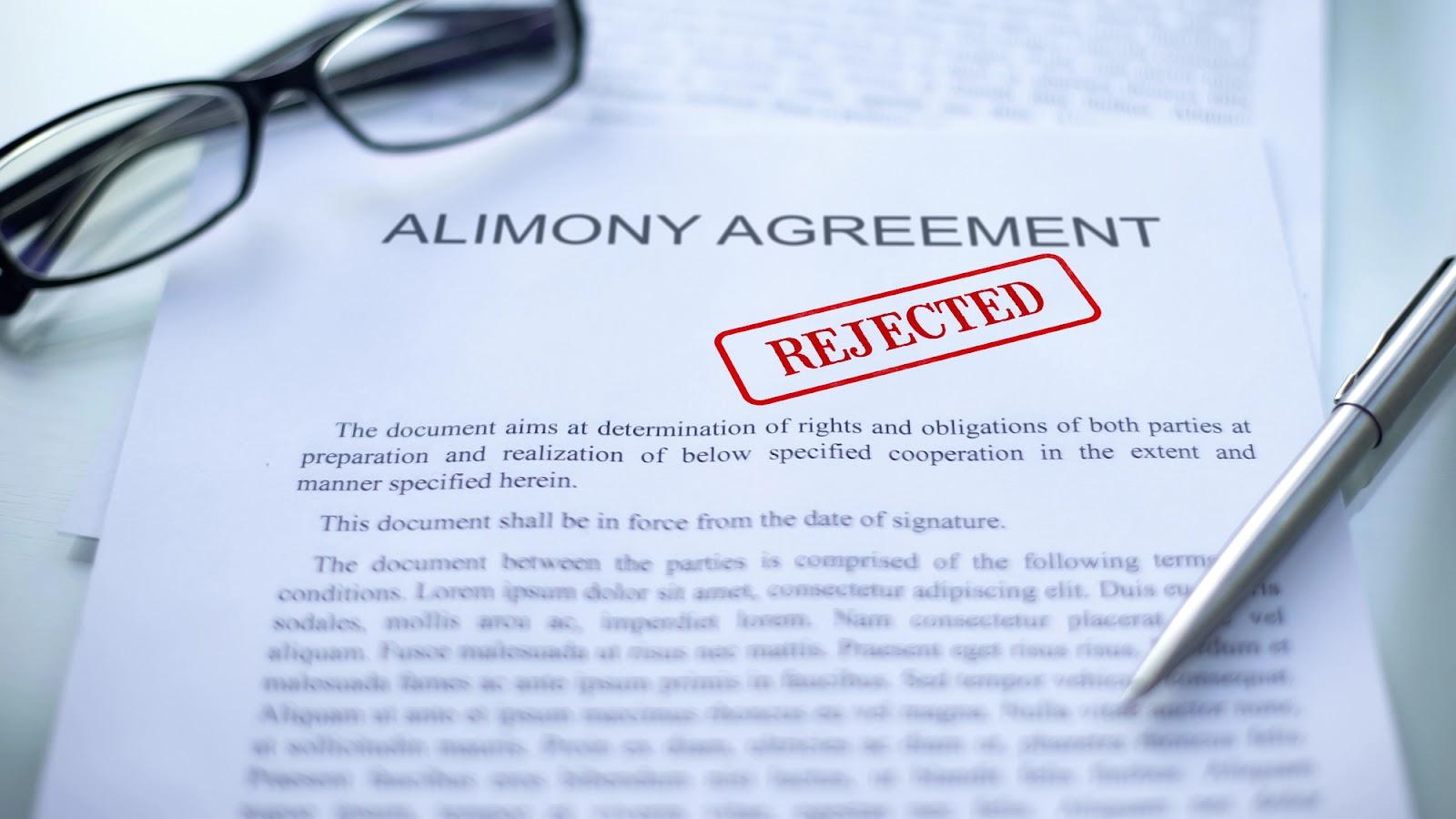 A pen and pair of glasses sit on the edge of a piece of paper titled Alimony Agreement with a red Rejected stamp on it.